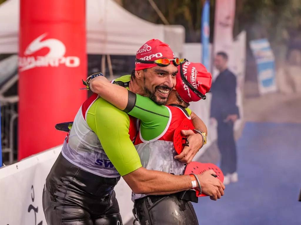 Two team-mates embrace at the finish, still in wetsuits with swim caps on (photograph from the 2025 edition): racing as a pair, the rules require partners to stay within 10 metres of each other in the water and within 50 metres on land.